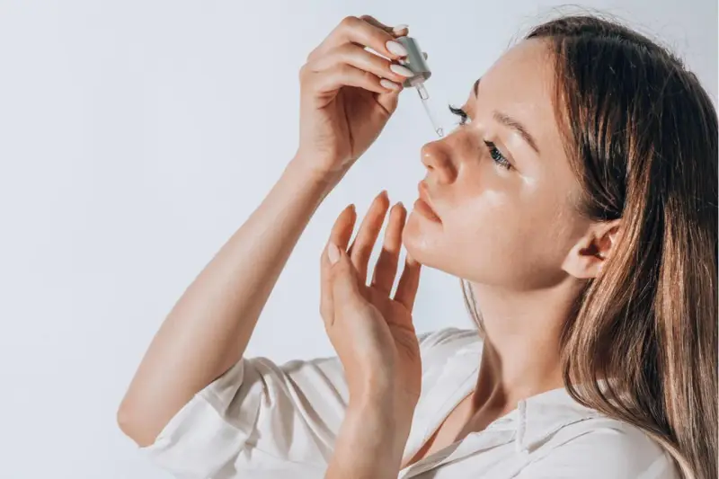 Frauenhand trägt Tropfen eines Serums auf ihr Gesicht auf. Ob Squalan oder Hyaluron, die Haut strahlt Feuchtigkeit aus.