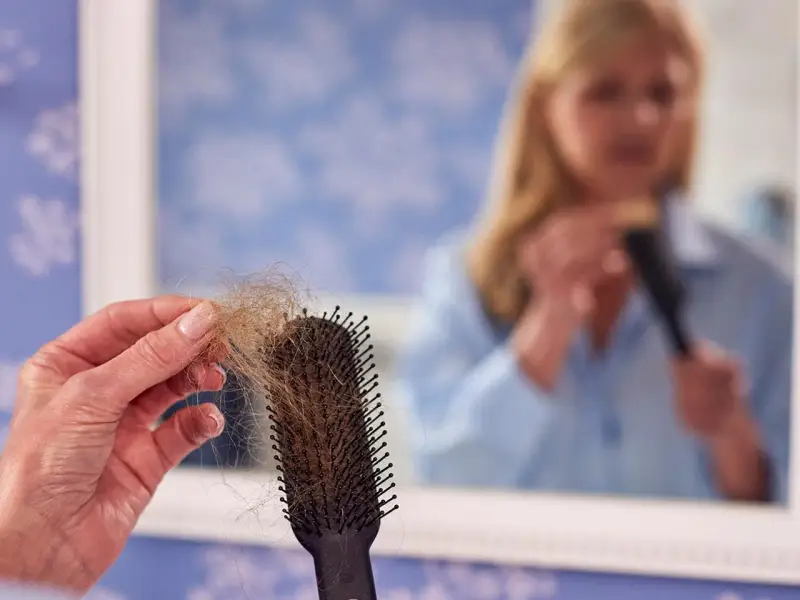 Frauen mit Haarausfall halten eine Bürste mit vielen Haaren. Im Spiegel sieht man eine Frau, die sich die Haare kämmt.